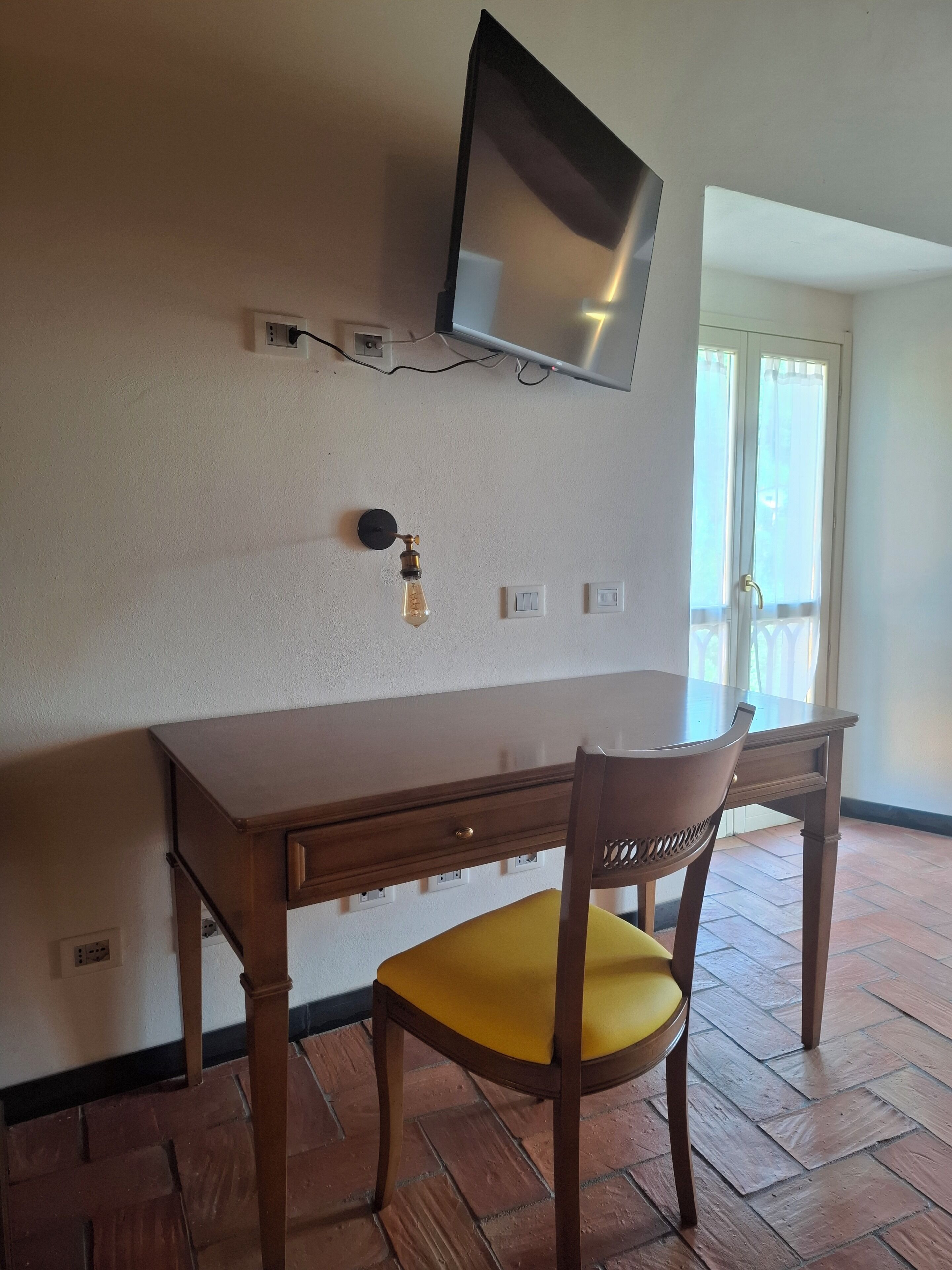 Una habitación con un escritorio de madera, una silla con cojín amarillo, un televisor montado y una ventana con cortinas.