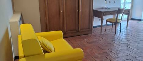 A room with a yellow sofa, a wooden wardrobe, a dining table with chairs, and a tiled floor.
