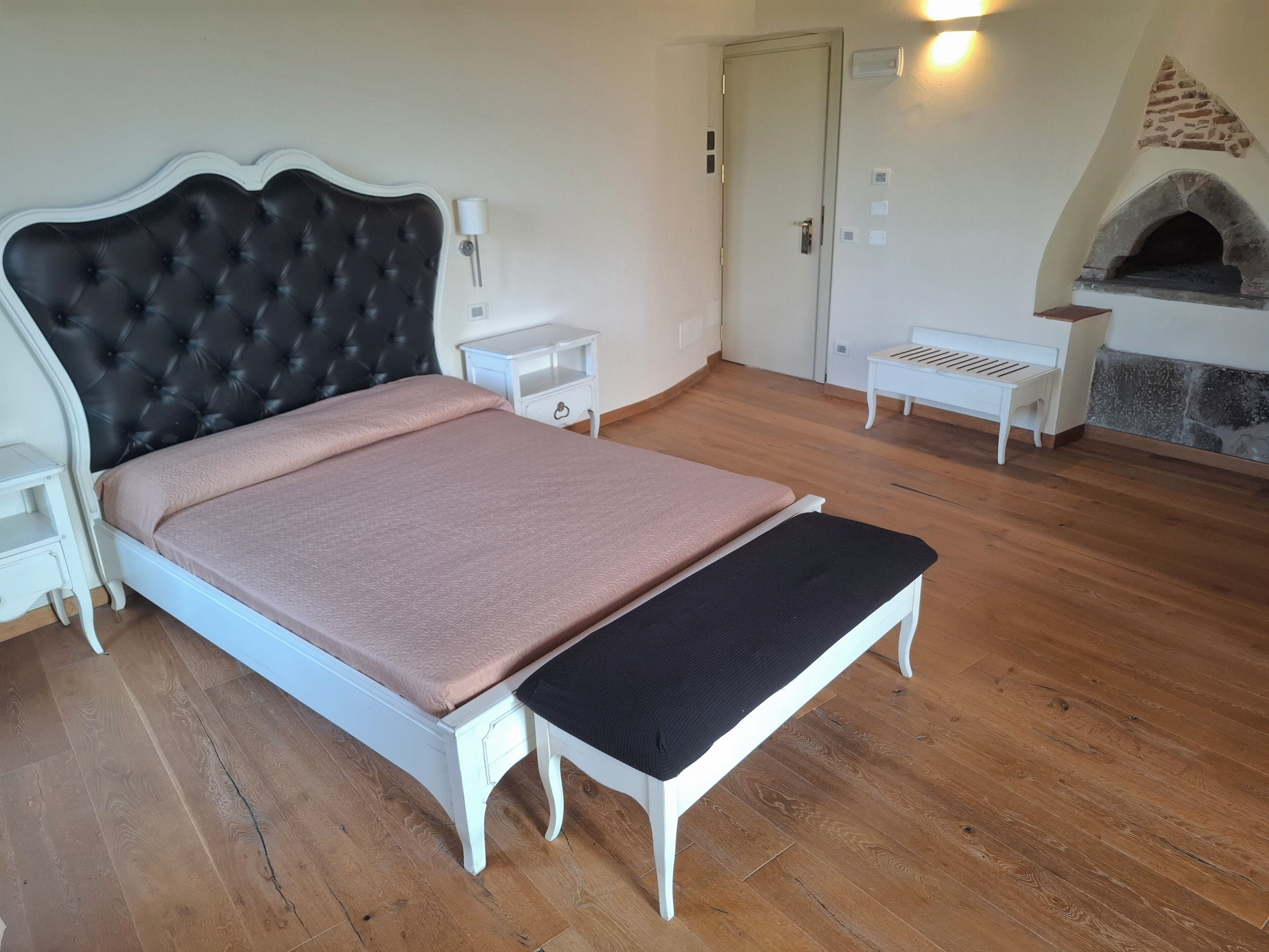 A bedroom with a bed, bedside tables, a bench, a fireplace, and a door.