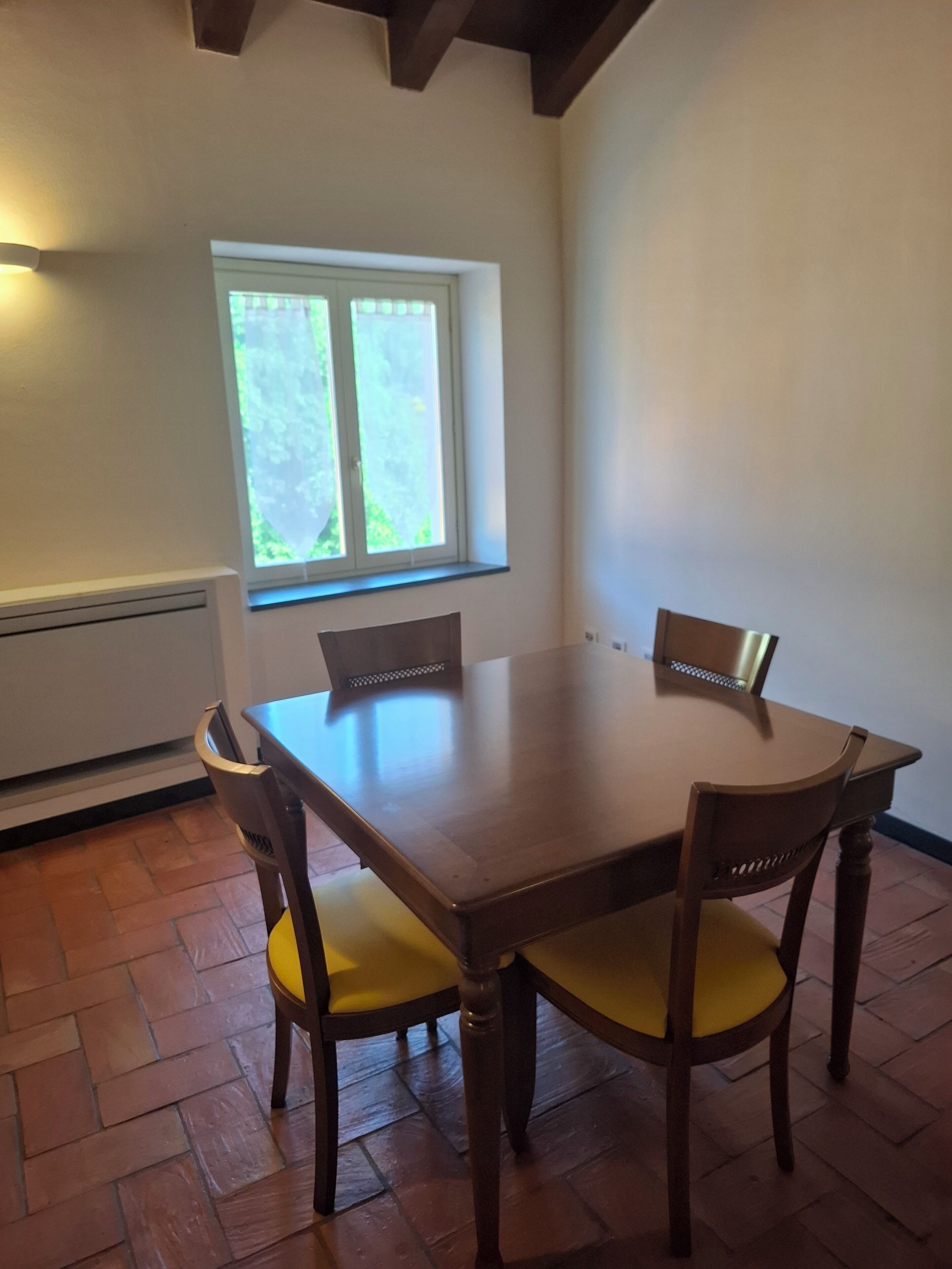 A dining table with four chairs, a window with a view of greenery, and a radiator on the wall.