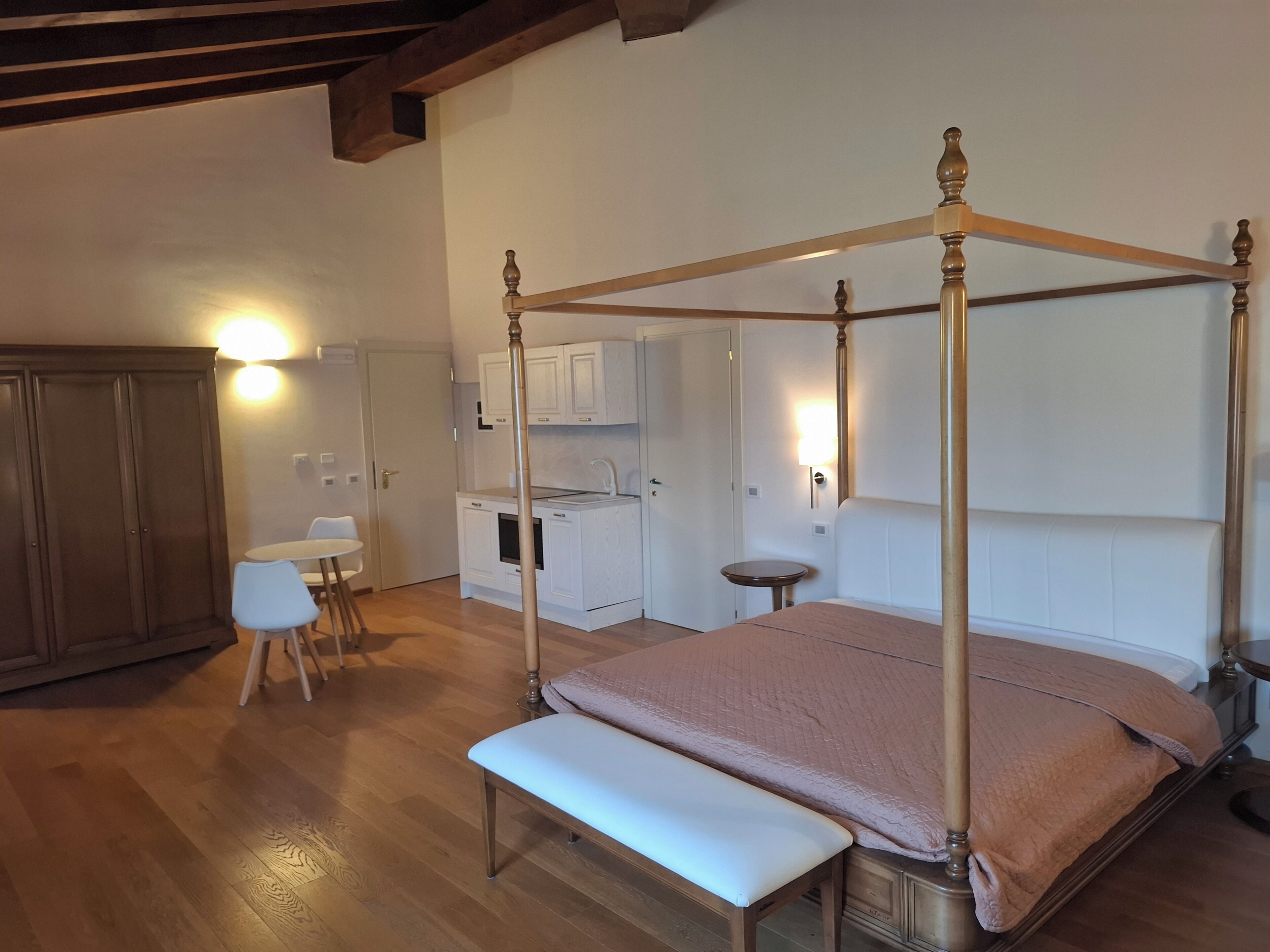 A bedroom with a four-poster bed, a small bedside table, a bench, a desk, and a chair. There is a kitchenette in the background.