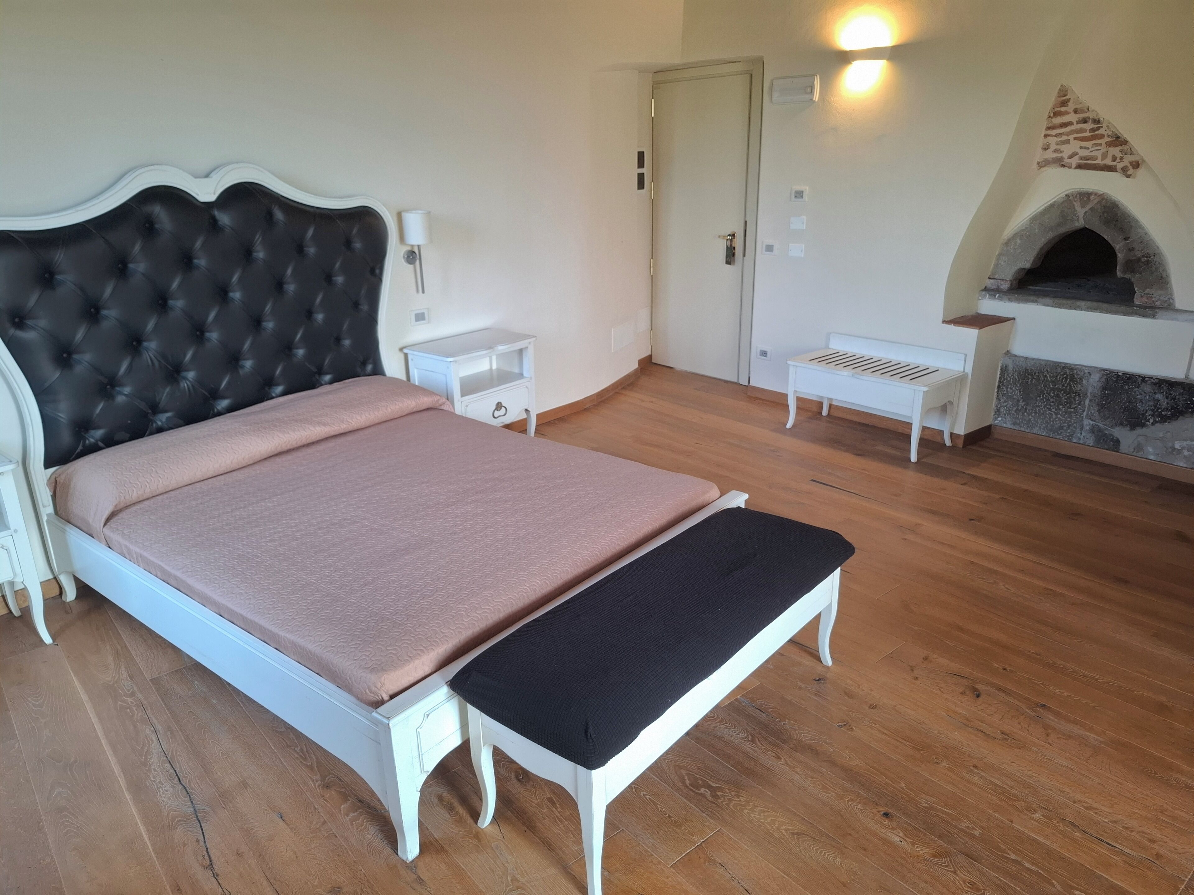 Een slaapkamer met een groot bed, een bankje, een nachtkastje, een open haard en een deur.