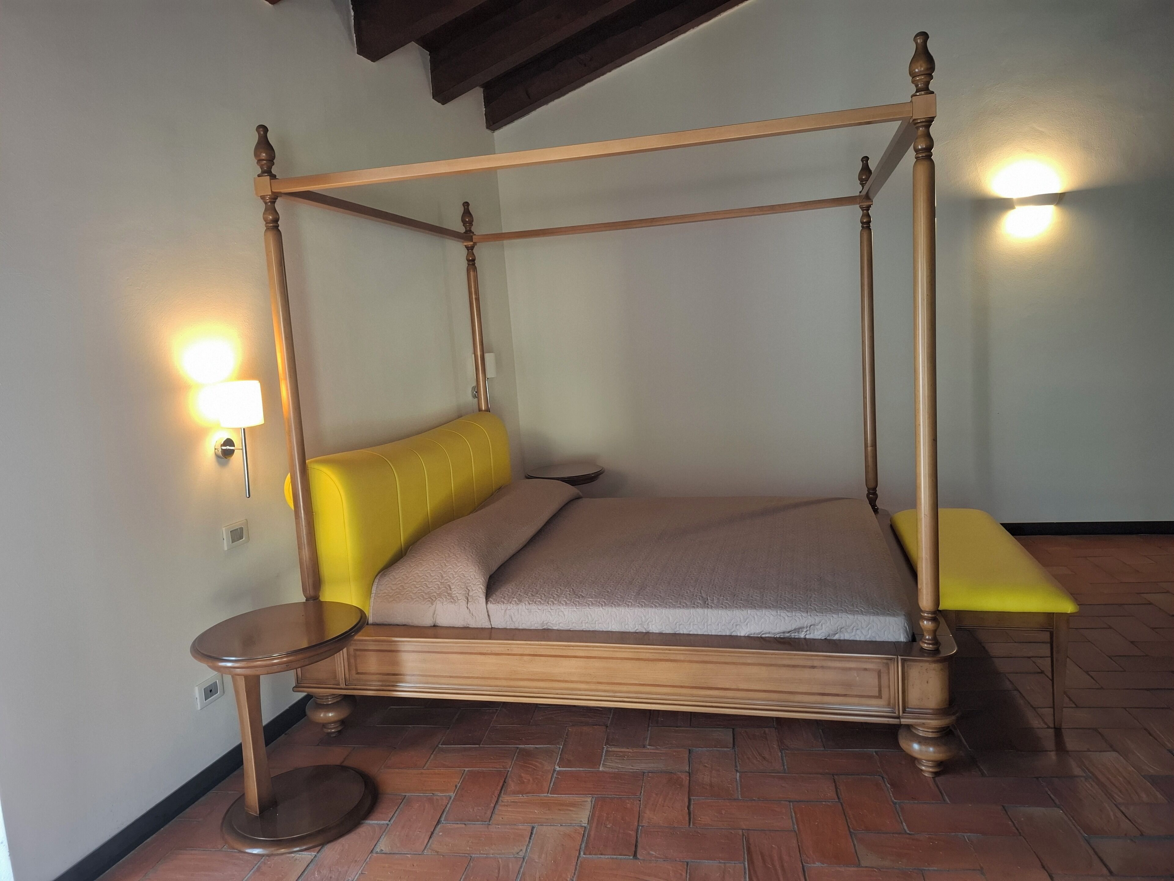 Una cama con dosel de madera y un cabecero tapizado en amarillo, flanqueada por dos mesitas redondas, cada una con una lámpara.