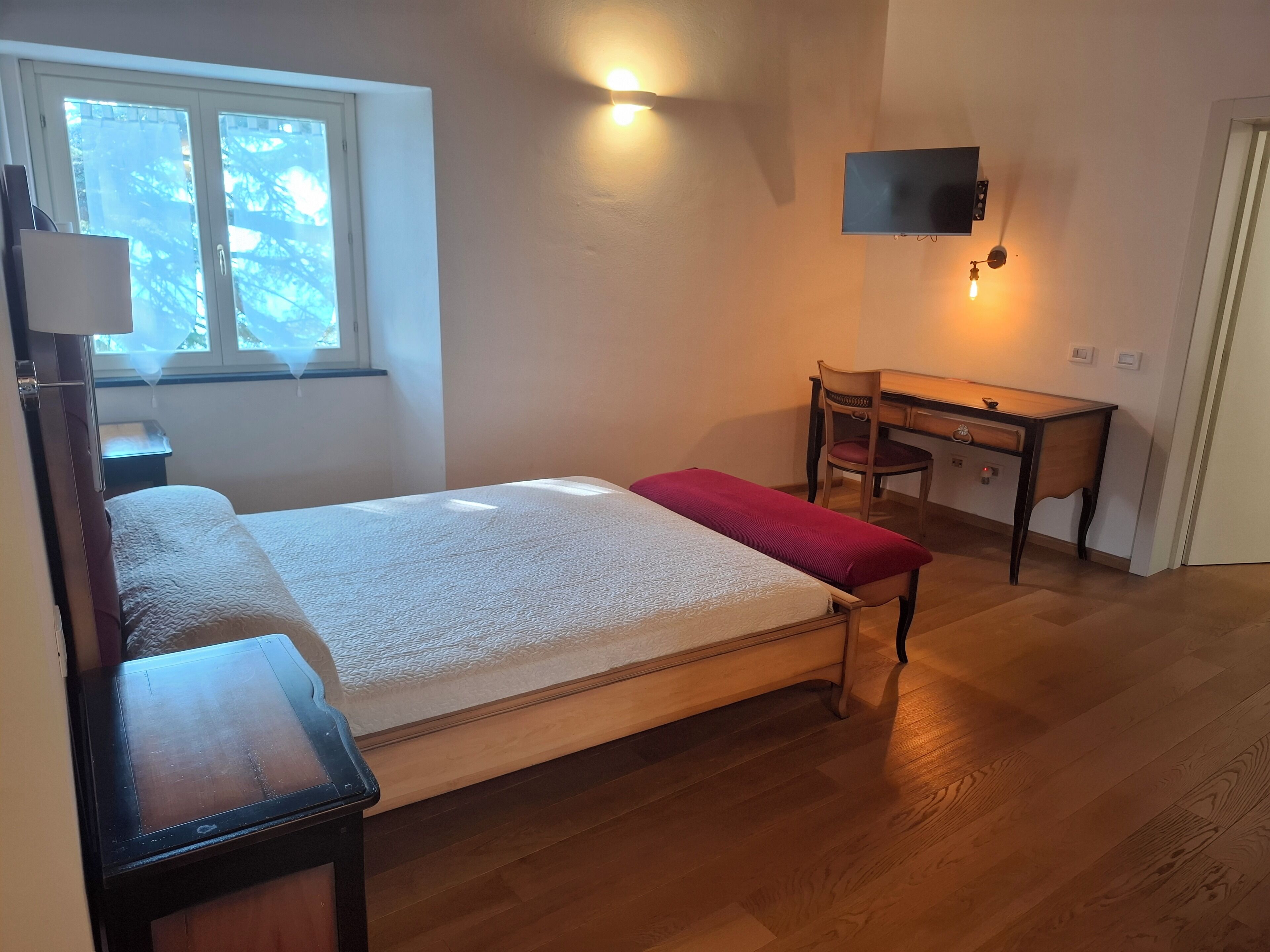 A bedroom with a bed, a desk, a chair, a TV, and a window.
