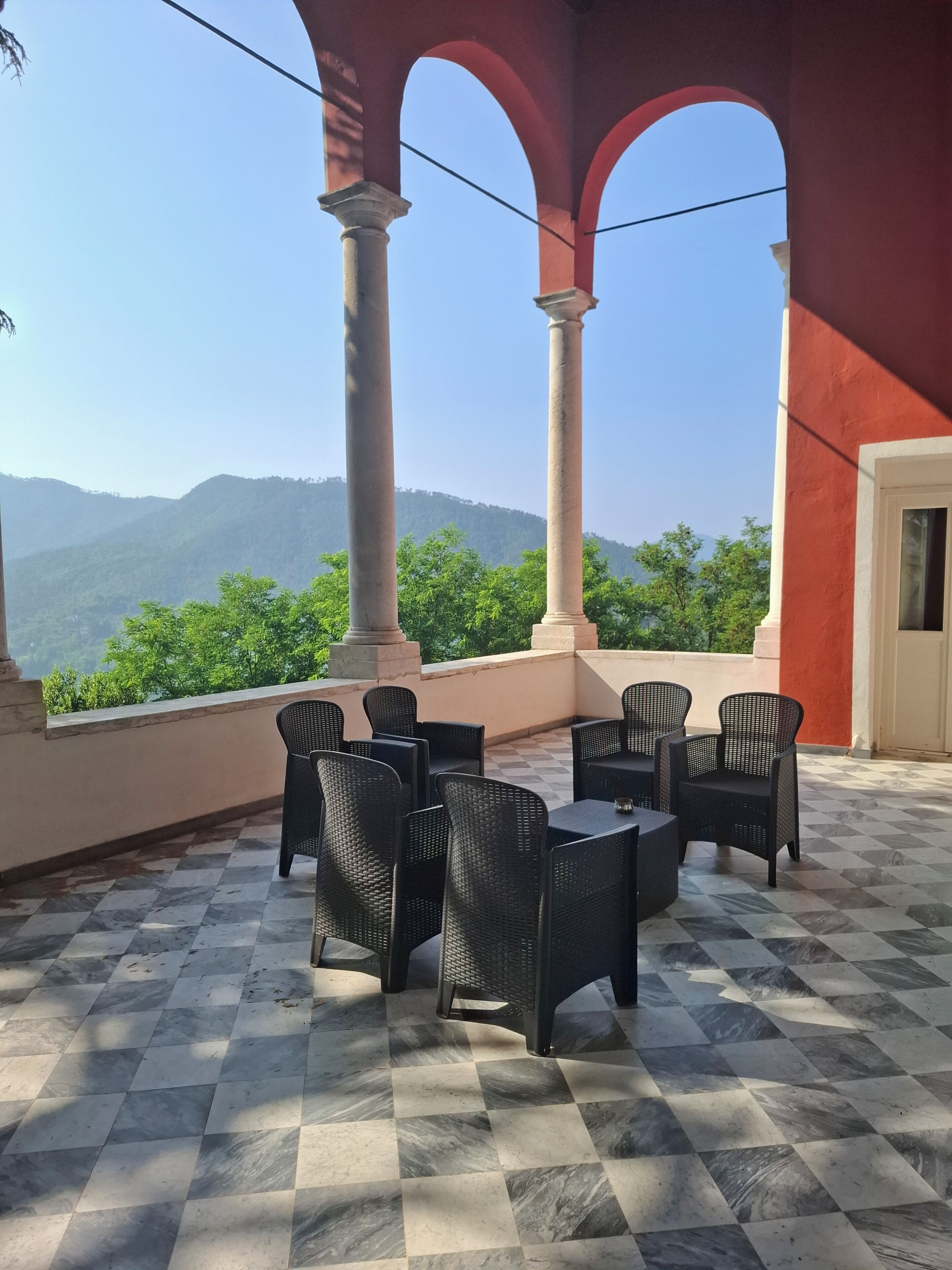 A terrace with checkered flooring, black wicker furniture, and a view of green hills under a clear sky.