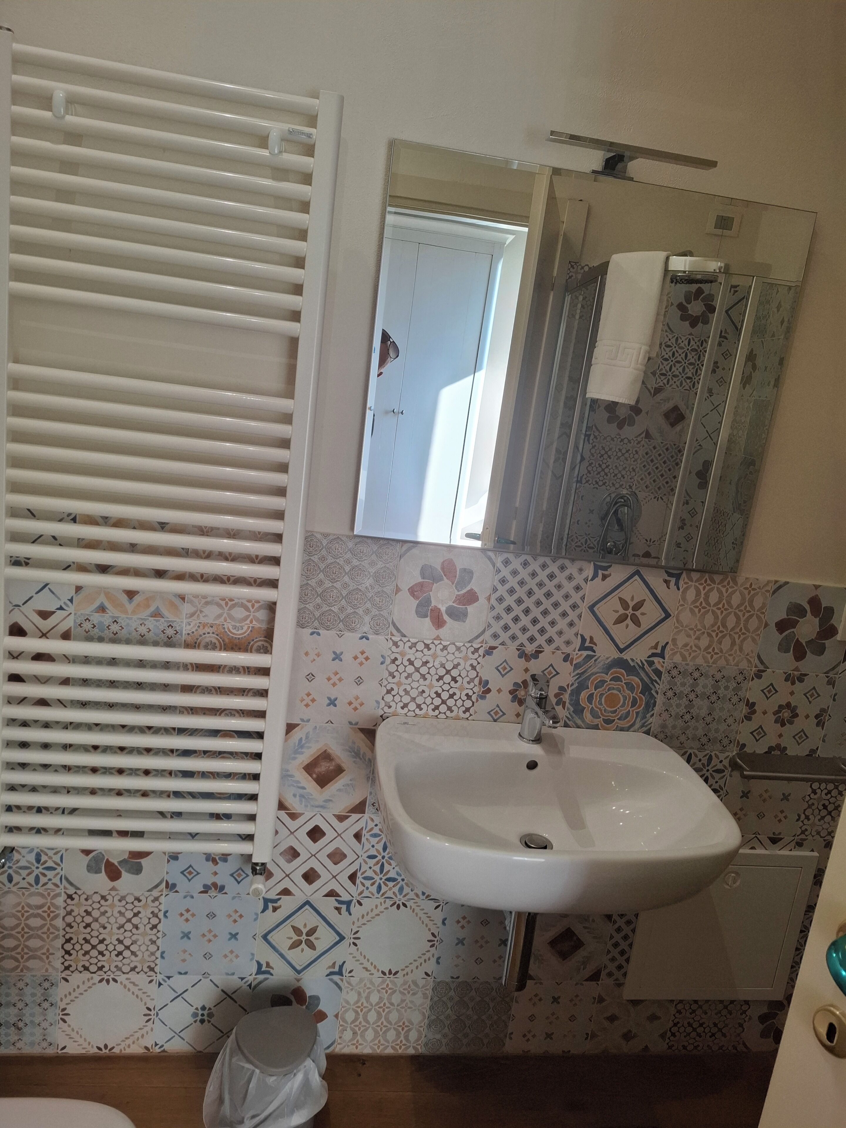 Un bagno con un lavandino bianco, piastrelle decorate, una finestra con persiane e uno specchio con un asciugamano.