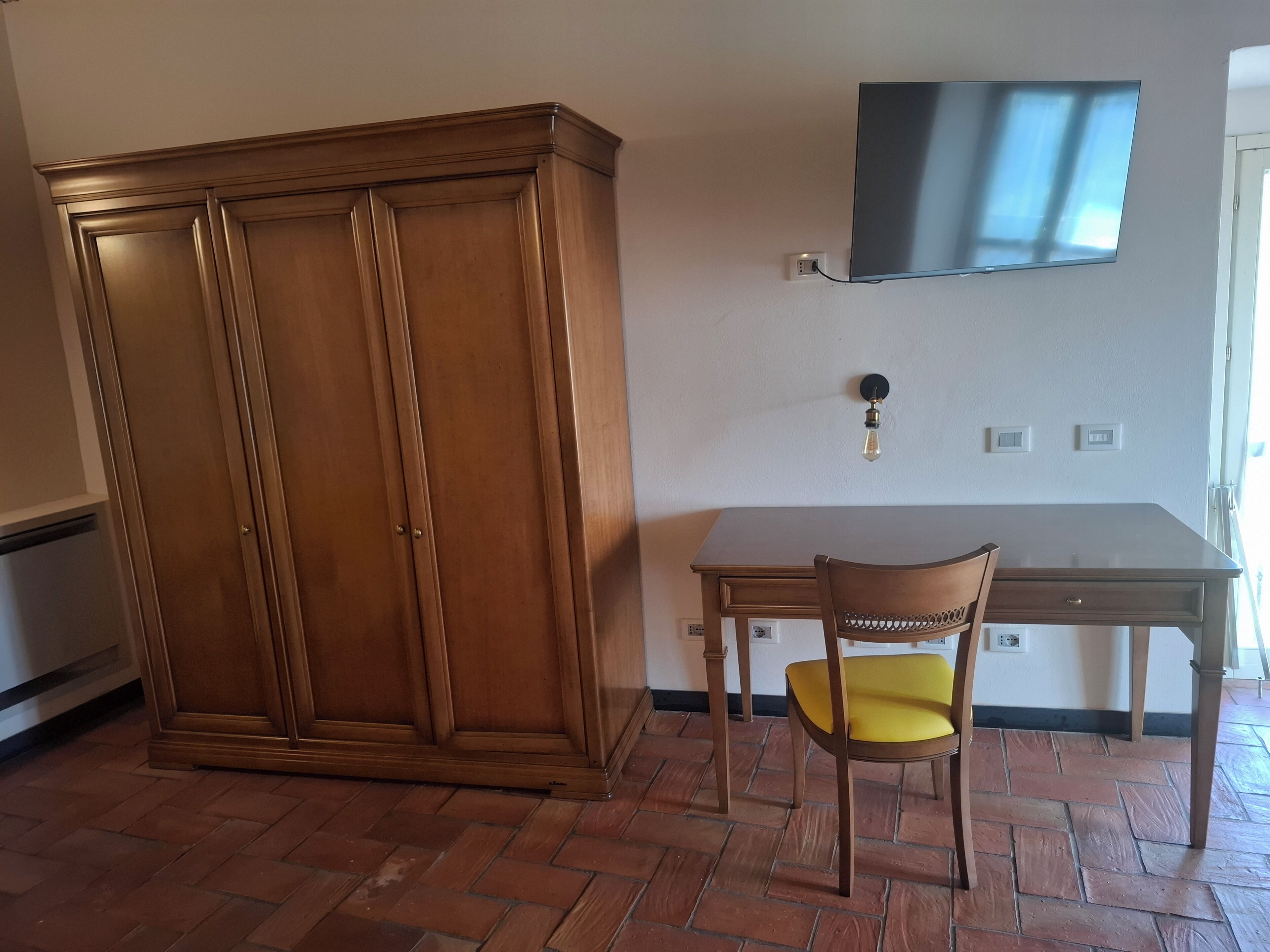 Ein Zimmer mit einem Holzschrank, einem Schreibtisch, einem Stuhl und einem an der Wand befestigten Flachbildfernseher.