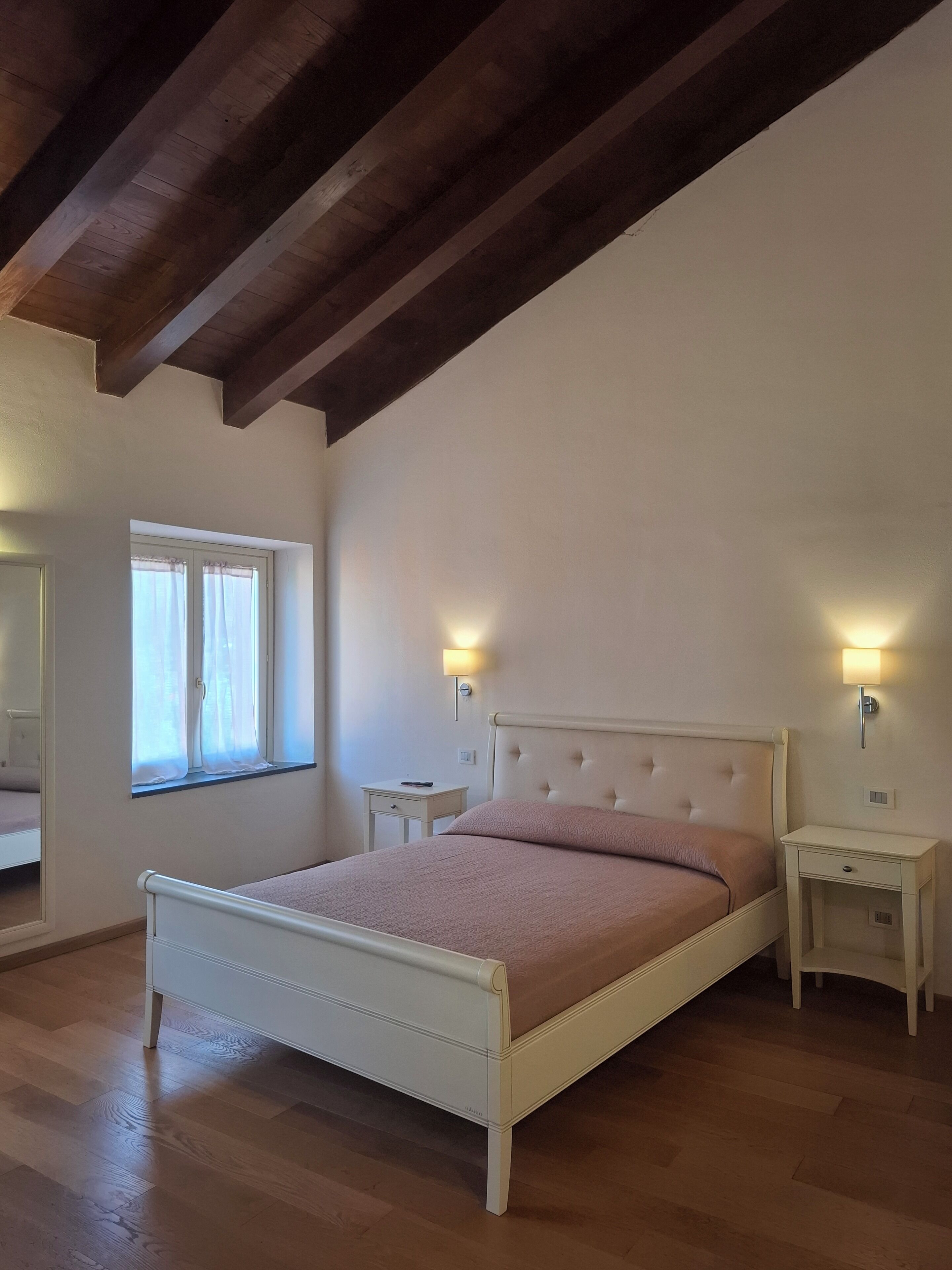 Ein Schlafzimmer mit einem Bett, Nachttischen, einem Fenster mit Vorhängen und Holzbalken an der Decke.