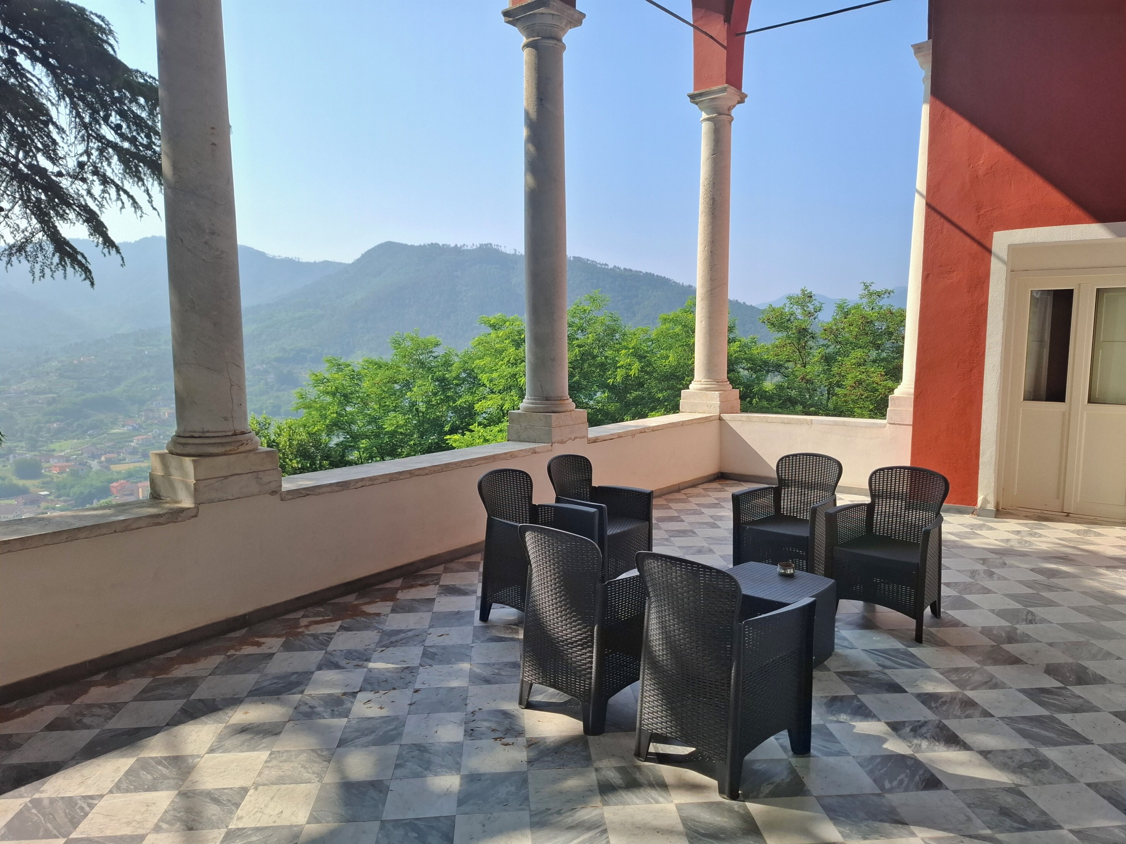 A terrace with checkered flooring, black wicker furniture, and a view of green hills and mountains.