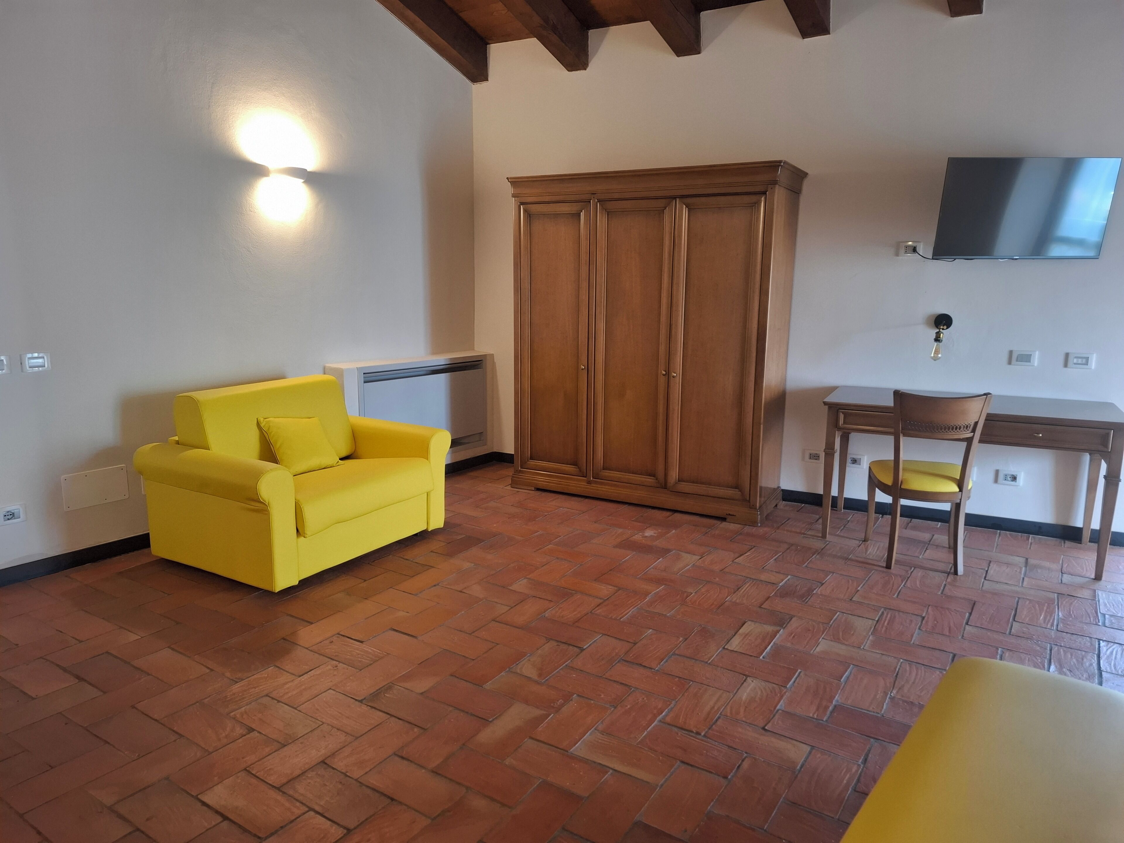 Una habitación con un sofá amarillo, un armario de madera, un escritorio con una silla y un televisor montado en la pared.