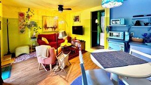A living room with a red sofa, a coffee table, and a dining table with chairs. There is a television, a lamp, and a bookshelf. - Disco Den on Toad Road (Plainfield)