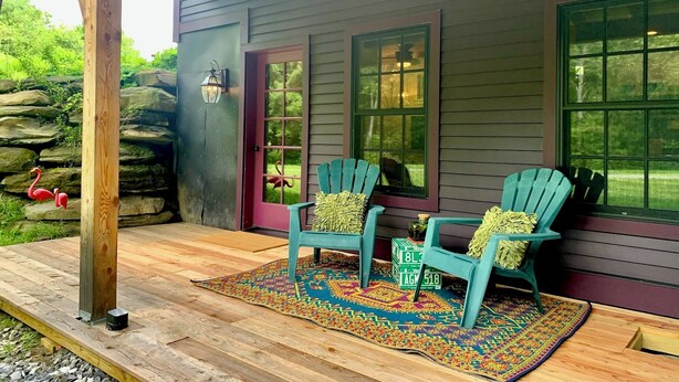 A wooden porch with two turquoise Adirondack chairs, a patterned rug, and a stone wall backdrop. - Disco Den on Toad Road (Plainfield)