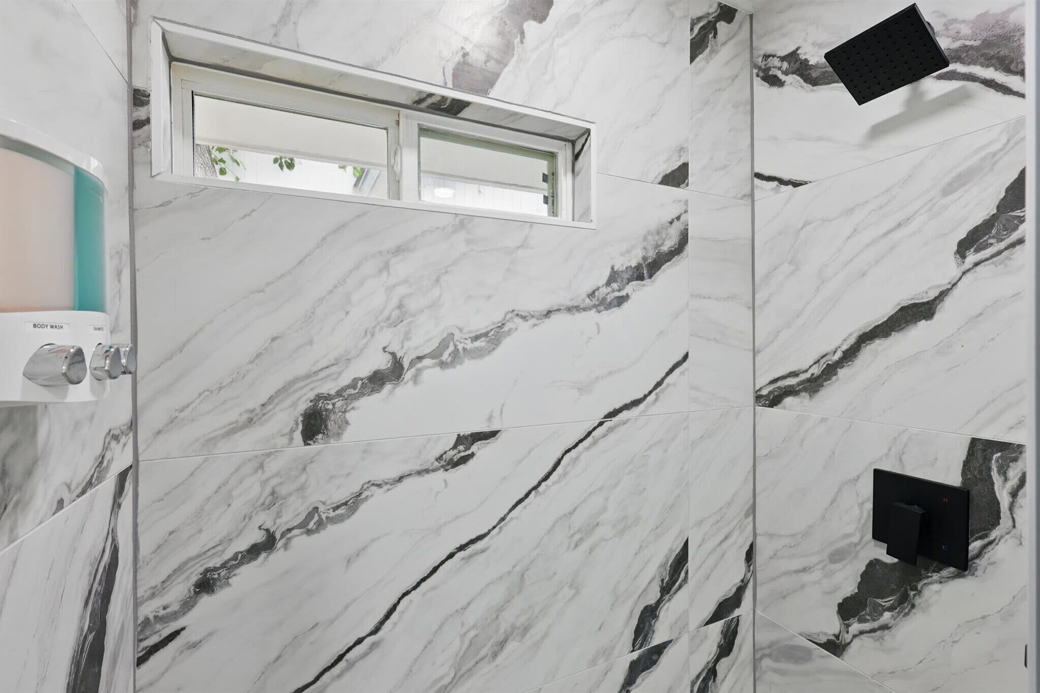 Un baño con paredes de mármol, una ventana, ducha y dispensador de jabón.