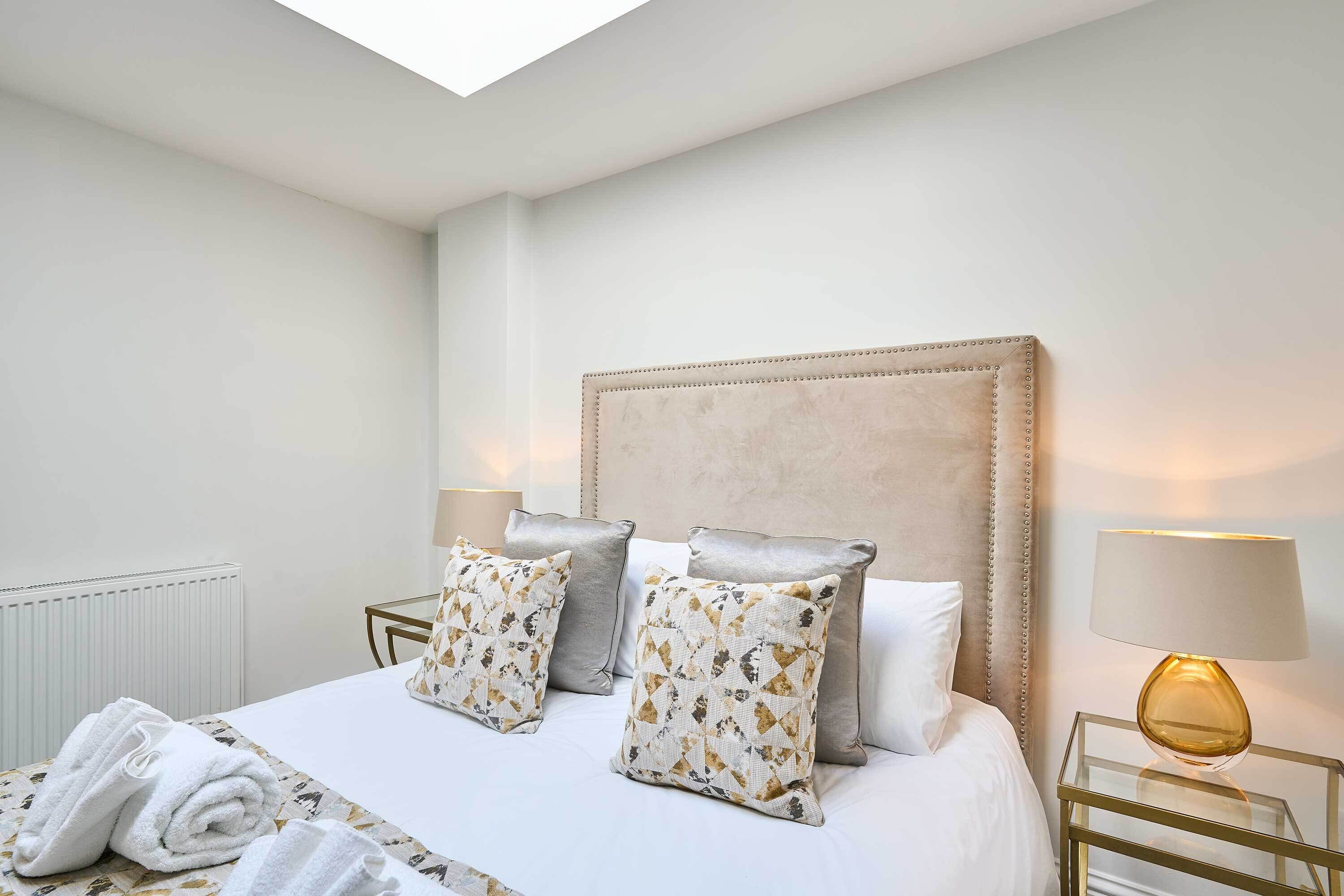A neatly made bed with patterned pillows, a bedside lamp, and a rolled towel on the floor.