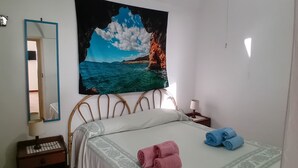 Une chambre à coucher comprenant un lit, une table de chevet, une lampe, un miroir et un tableau représentant un paysage côtier.