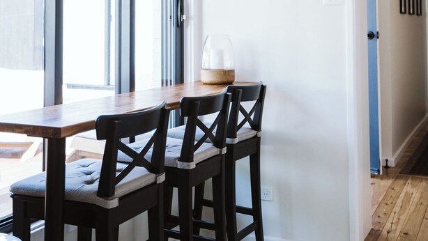 Un comedor con una mesa de madera y cuatro sillas, un letrero en la pared que dice "BEACH" y vistas a través de grandes ventanales.