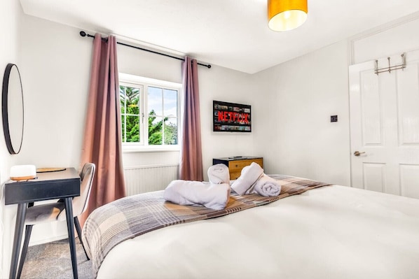 A hotel room with a bed, a desk, a chair, a window with curtains, a wall-mounted TV, and a door.