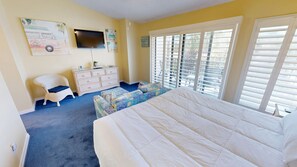 Une chambre avec un grand lit, un téléviseur fixé au mur, une commode, une chaise et une fenêtre avec des stores.
