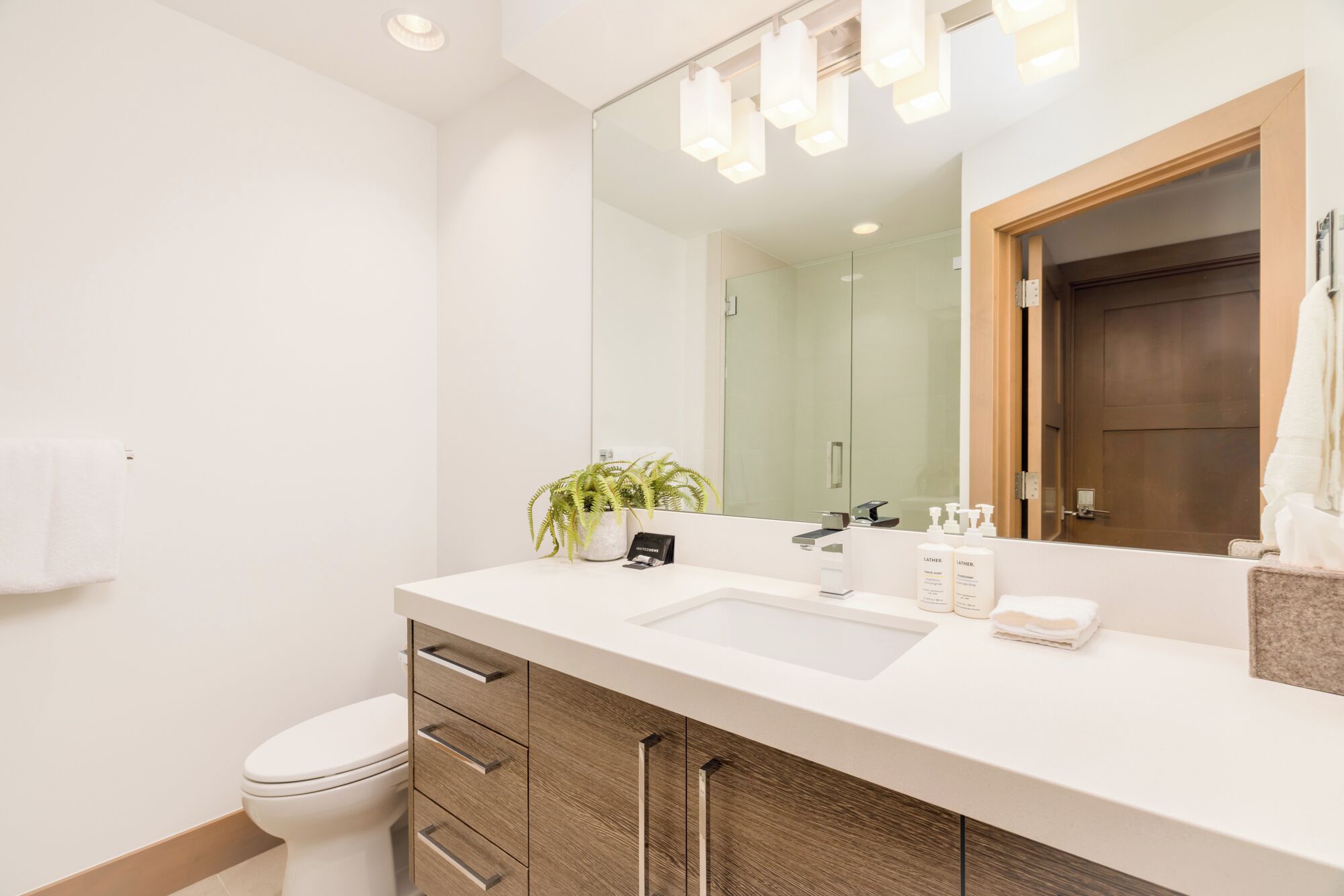 A modern bathroom with a white sink, wooden cabinets, a mirror, and a potted plant.