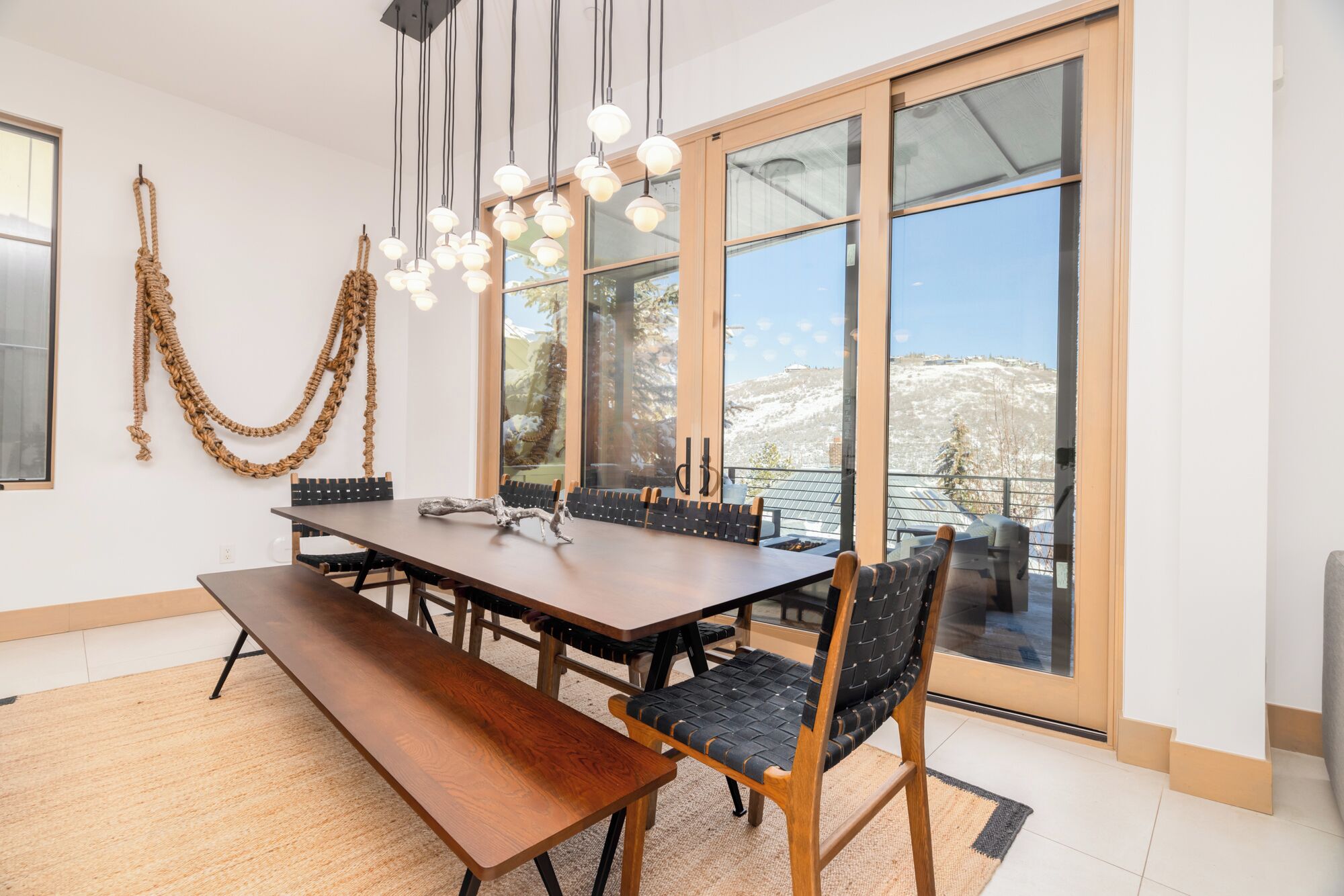 A dining area with a large table, chairs, and a bench. There are hanging lights and a view of a snowy landscape through large windows.