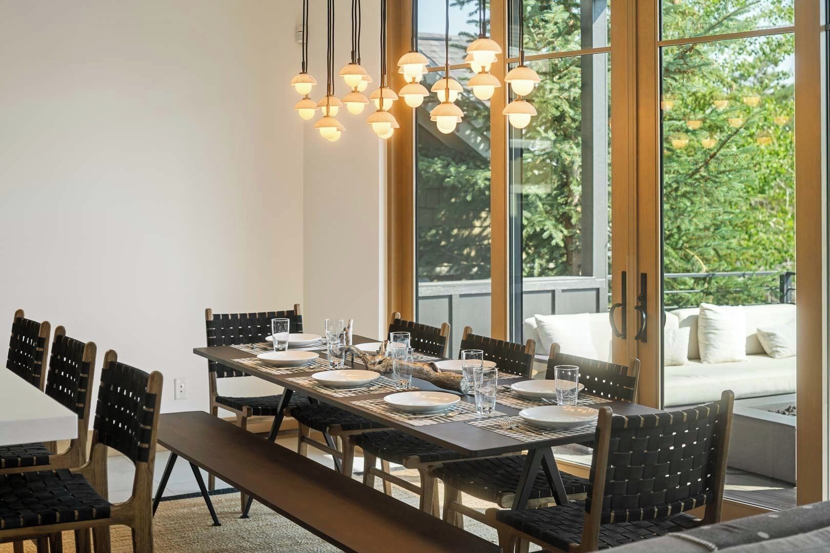 A dining area with a long table set for six, surrounded by chairs, with a large window offering a view of greenery outside.