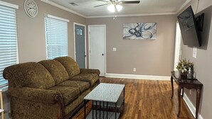 Un salon avec un canapé marron, une table basse, un plancher en bois, un ventilateur de plafond, une horloge, un tableau et un téléviseur fixé au mur.