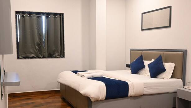 Une chambre à coucher avec un lit, un plancher en bois, une fenêtre avec des rideaux, une table de chevet et un luminaire fixé au mur.