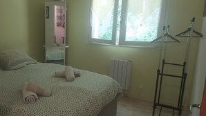 A neatly made bed with rolled towels, a radiator, a window with a view of greenery, and a clothing rack. - Country apartment close to the forest. You can go for a nice walk, (Boussens)