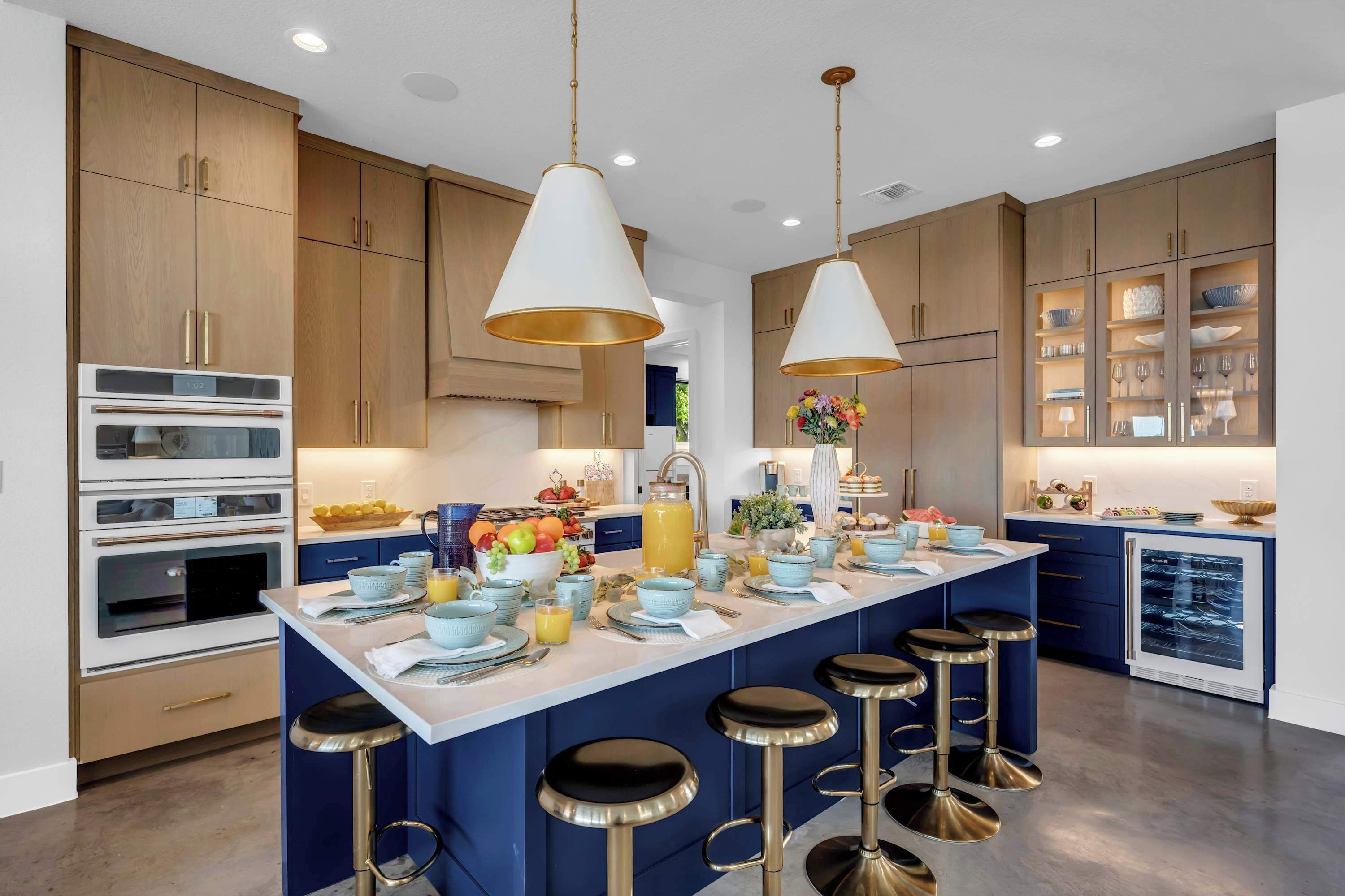 A modern kitchen with a central island, bar stools, built-in ovens, and wooden cabinets.