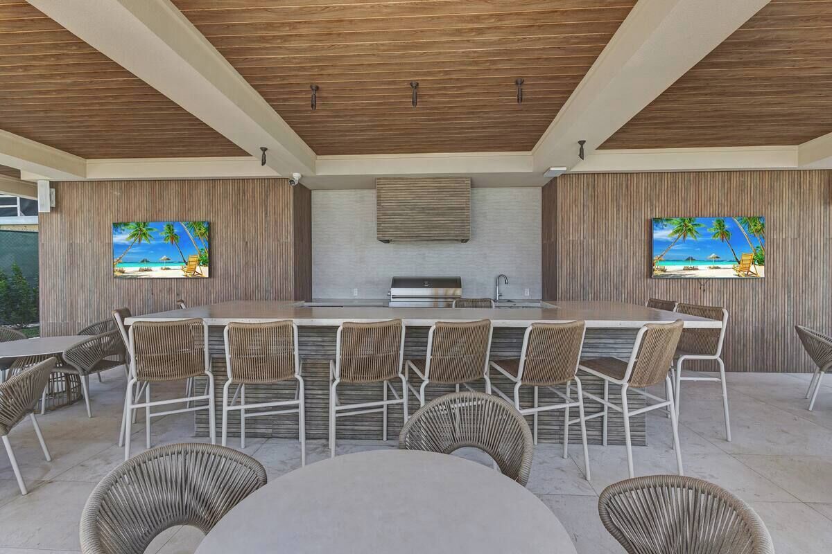 An outdoor bar area with a long counter, wicker chairs, and two TV screens displaying beach scenes.