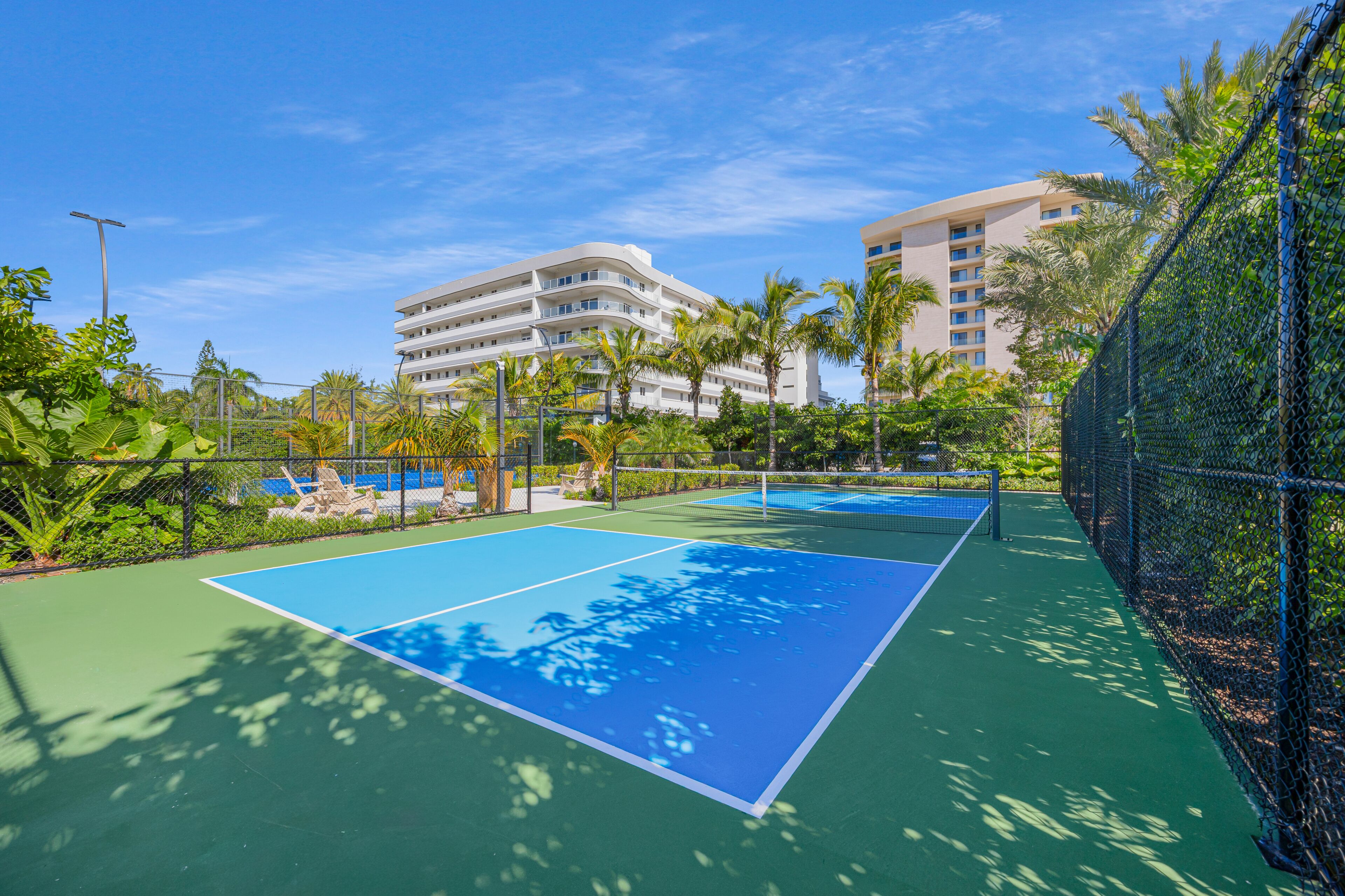 A tennis court with a net, surrounded by greenery and palm trees, with a building in the background.