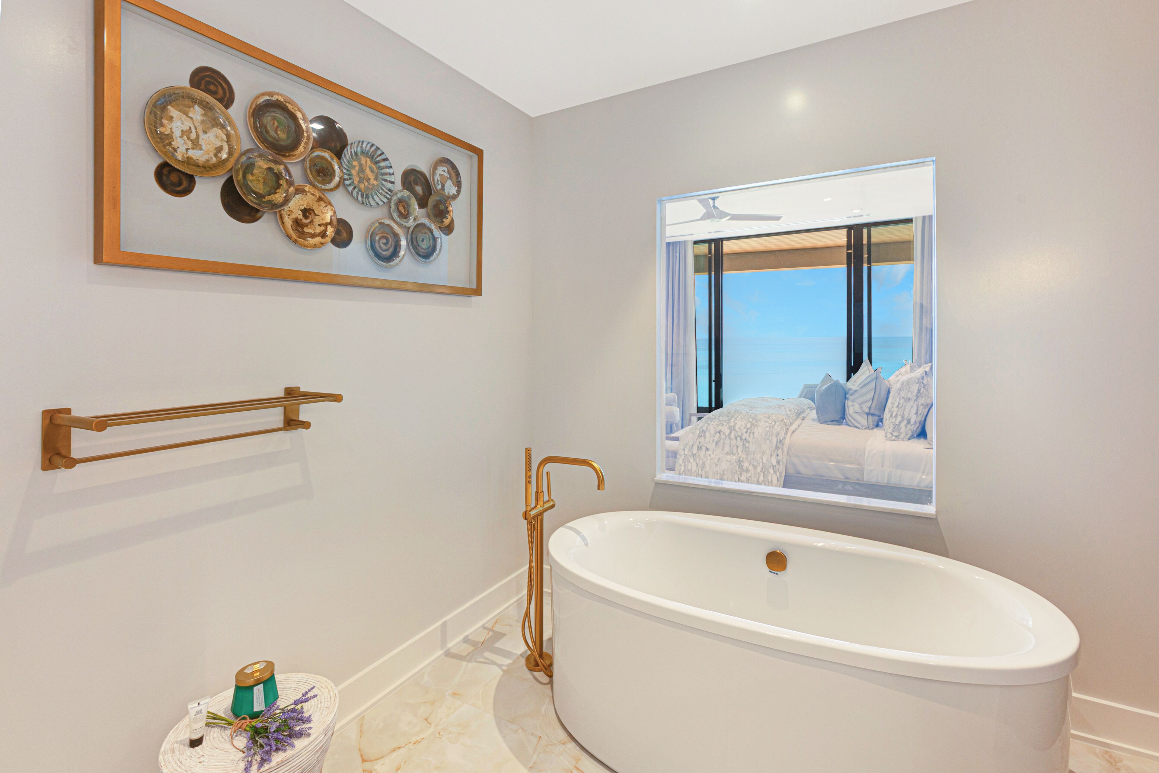 A modern bathroom with a freestanding bathtub, a framed artwork above the tub, a towel rack, and a window with a view of a bedroom.