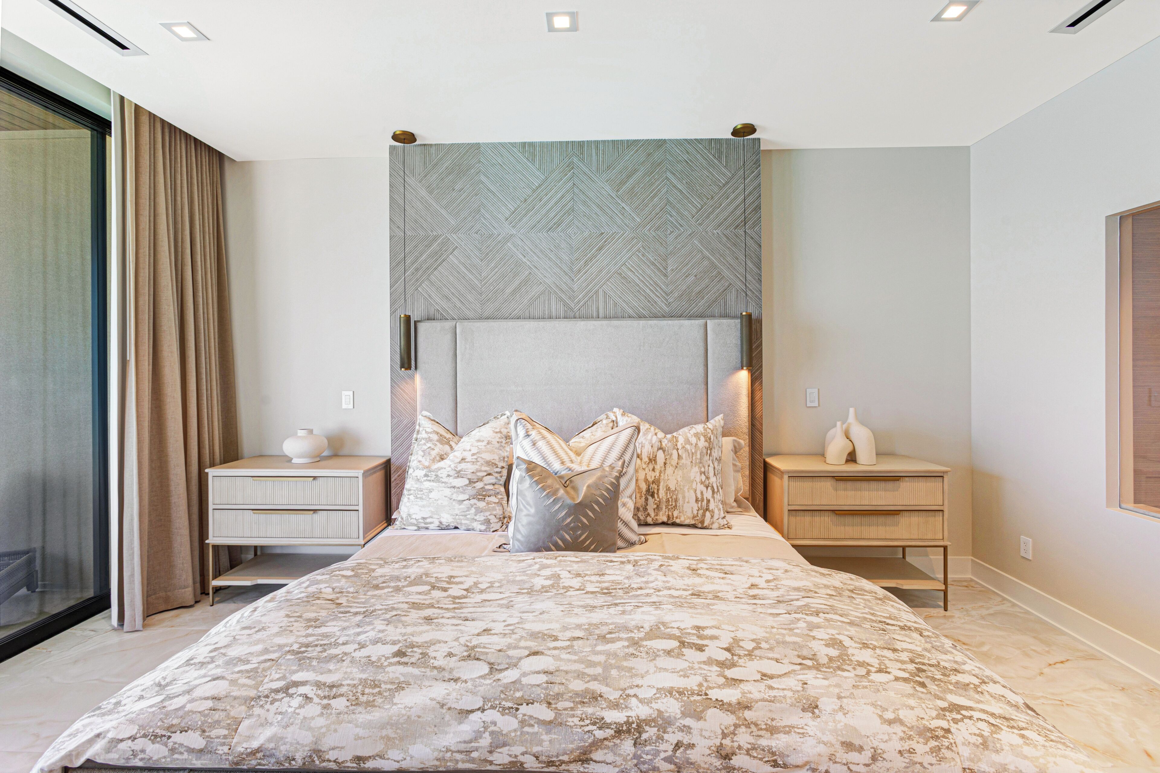 A bedroom with a large bed, two bedside tables, a headboard with a geometric pattern, and a large window with curtains.