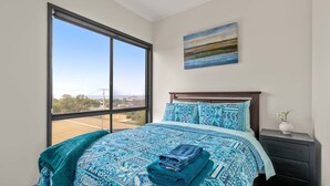 A bedroom with a large window, a bed with patterned bedding, a nightstand, and a painting on the wall.