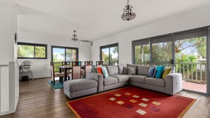 A spacious living room with a grey sectional sofa, a dining area with a table and chairs, and large windows offering a view of the outdoors.