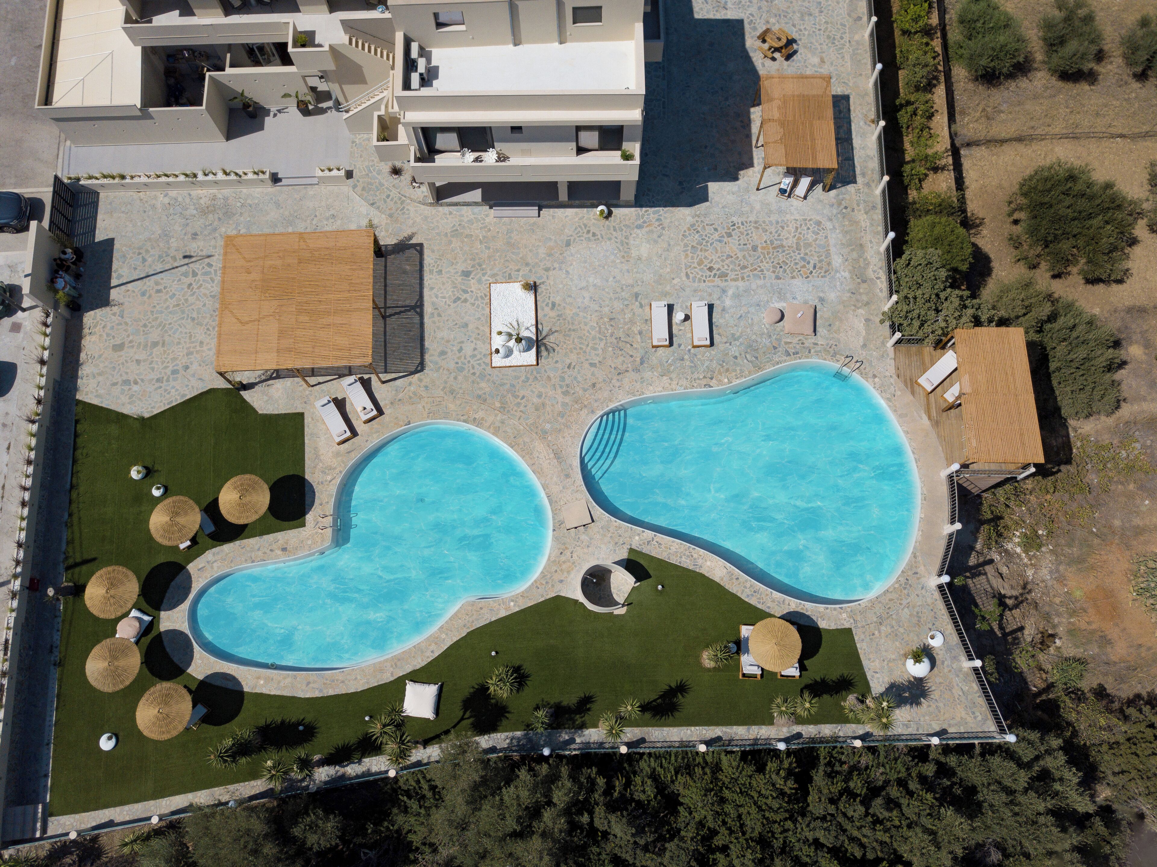 An aerial view of a resort with two swimming pools, sun loungers, umbrellas, and a small building.