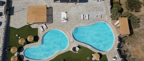 An aerial view of a resort with two swimming pools, sun loungers, umbrellas, and a small building.