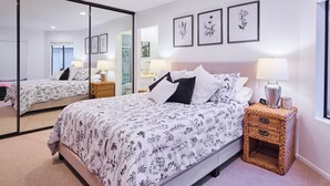 A bedroom with a large mirror, a bed with floral bedding, two bedside tables with lamps, and framed botanical prints on the wall.