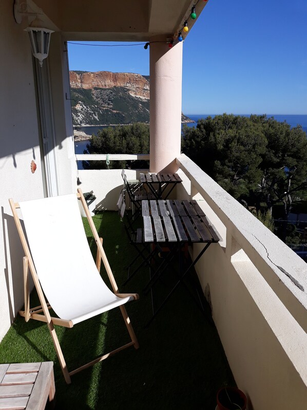 Un balcone con una sedia bianca, un tavolo di legno e vista su un paesaggio montuoso e il mare.