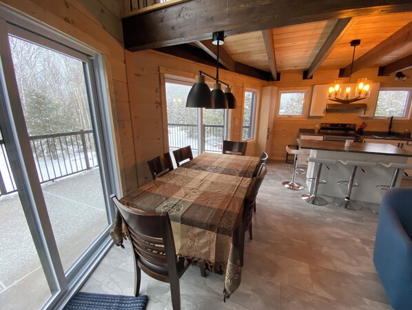 A spacious dining area with a large table and chairs, a kitchen with a bar, and large windows offering views of the outdoors.