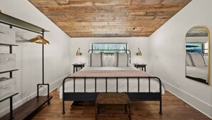 A bedroom with a wooden ceiling, a bed with a metal frame, bedside tables with lamps, a mirror on the wall, and a window with a view.