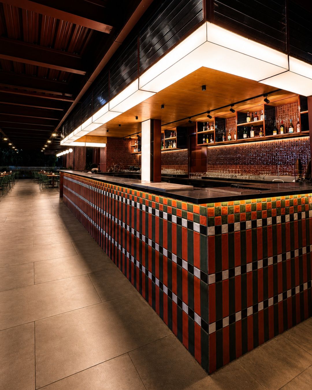 Uma área de bar com balcão revestido de azulejos, prateleiras com garrafas e teto com vigas aparentes.