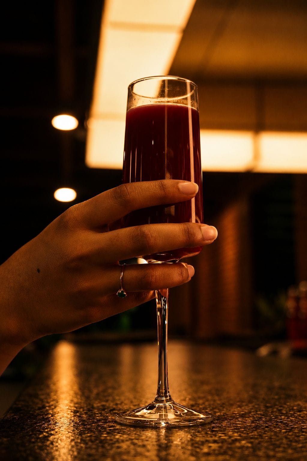 Una mano sosteniendo un vaso alto con un líquido rojo, posiblemente un cóctel, sobre una encimera.