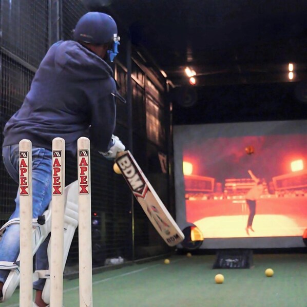 A person playing cricket in an indoor setting with a large screen displaying a virtual player. - Sleepover (Bengaluru)