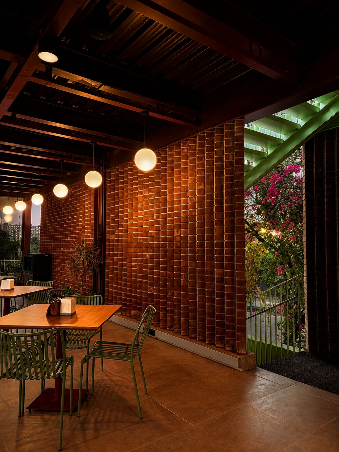 A restaurant interior with brick walls, hanging pendant lights, and a view of a green staircase and flowers.