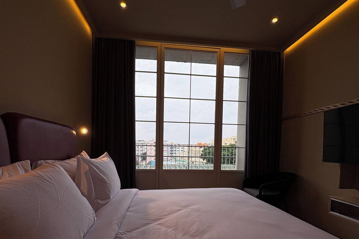 Ein Hotelzimmer mit einem großen Bett, einem Deckenventilator, einem an der Wand montierten Fernseher und einem Fenster mit Blick auf Gebäude.