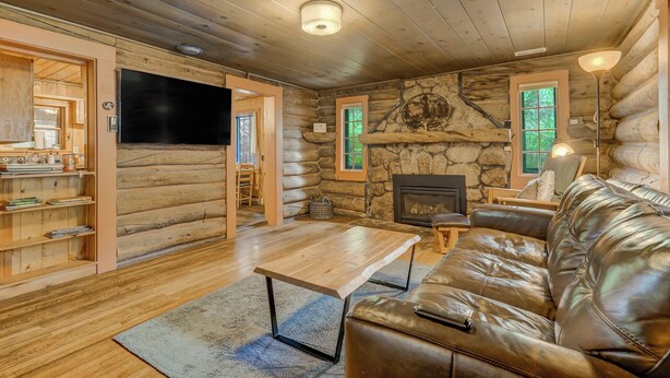 A log cabin interior with a wooden table, leather sofa, stone fireplace, and a flat-screen TV mounted on the wall.