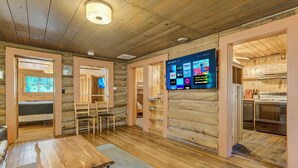 A log cabin interior with a wooden dining table, a TV mounted on the wall displaying various streaming services, and a kitchen area visible through an open doorway.