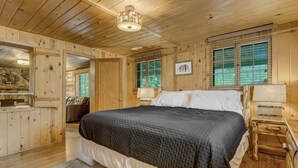 A wooden cabin bedroom with a large bed, a nightstand, a dresser, and a window with blinds.