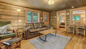 A log cabin interior with a leather sofa, wooden coffee table, and a dining area with chairs.